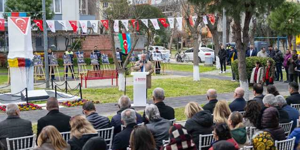 Bornova'da düzenlenen Hocalı Katliamı anma töreninde, kürsüde konuşma yapan bir yetkili ve onu dinleyen katılımcılar yer alıyor. Park alanında, üzerinde Türk ve Azerbaycan bayraklarının asılı olduğu bir anıtın çevresinde, Hocalı'yı anlatan fotoğrafların sergilendiği şövaleler bulunuyor. Katılımcılar açık havada kurulan sandalyelerde otururken, arka planda ağaçlar ve binalar görülüyor.