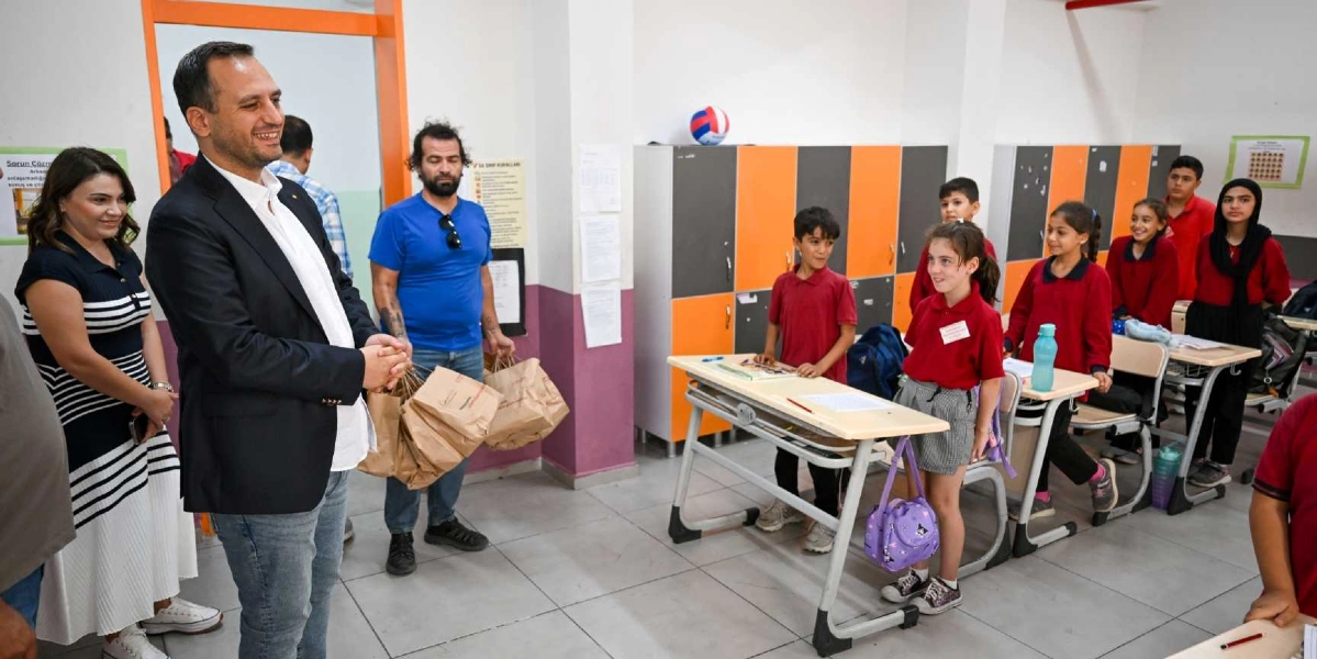Sınıf ortamında, ellerinde içinde yiyecek bulunan kağıt paketler taşıyan bir erkek ve yanında duran bir kadın, sıralarında oturan ilkokul öğrencilerine destek ziyareti gerçekleştiriyor. Öğrencilerin çoğu kırmızı okul formaları giymiş durumda ve sınıfta turuncu-gri dolaplar ile duvarlarda asılı bilgilendirme kağıtları bulunuyor.