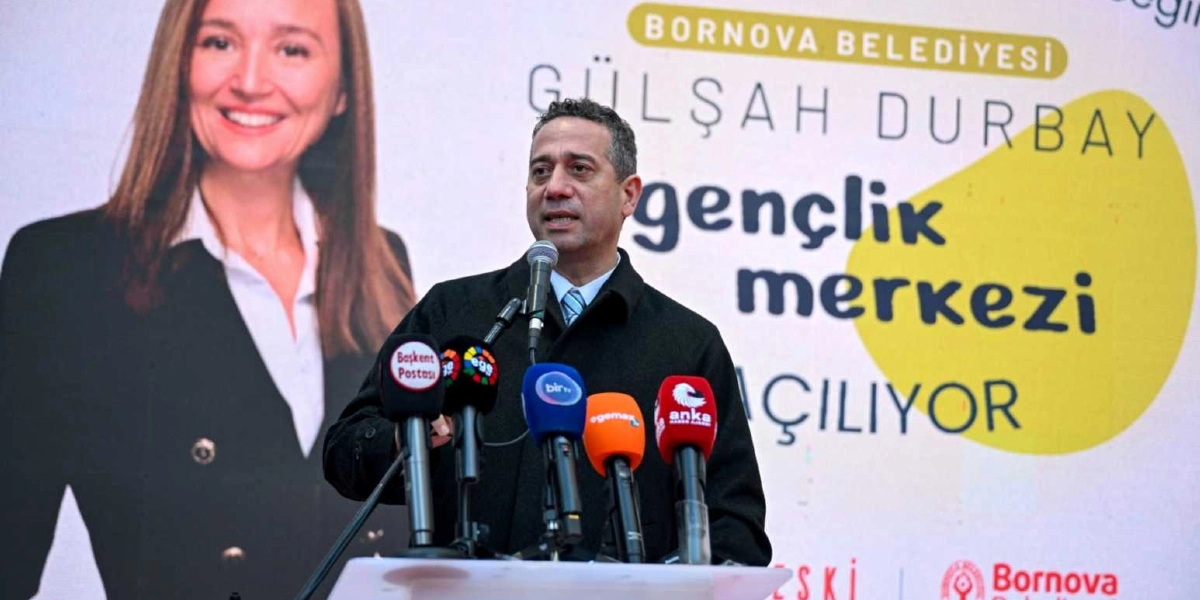 Bornova Belediyesi Gençlik Merkezi açılışında kürsüde konuşma yapan bir erkek, önündeki mikrofonlara hitap ediyor. Arka planda üzerinde Gülşah Durbay'ın fotoğrafının ve 