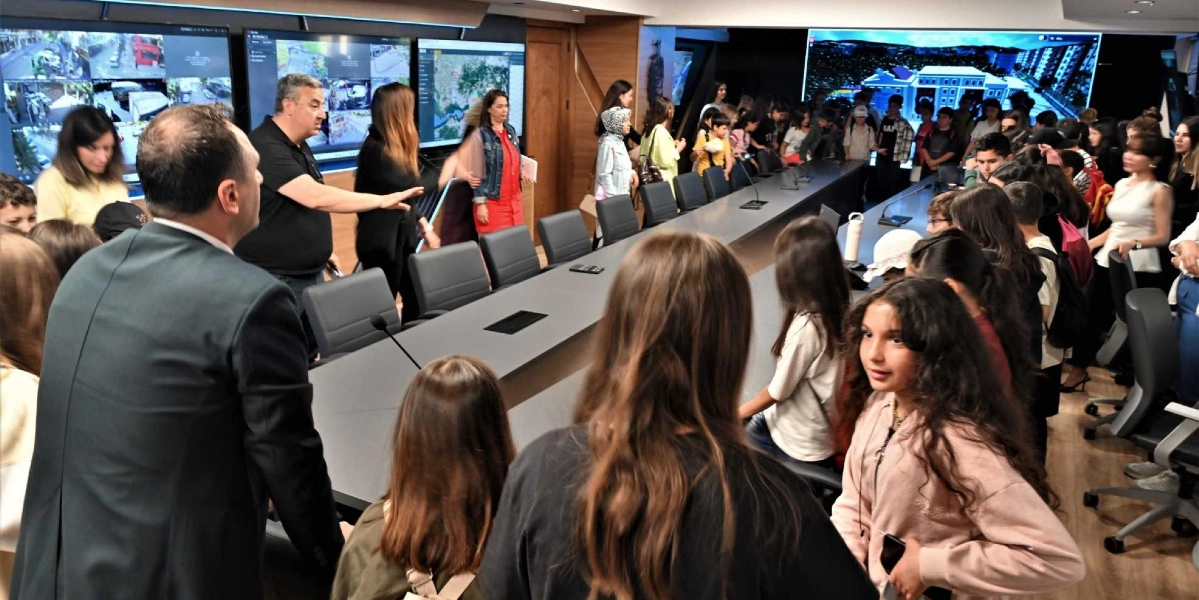 Bornova Belediyesi'nin modern bir toplantı salonunda, büyük ekranların önünde duran bir grup öğrenci ve onlara rehberlik eden yetkililer. Uzun bir toplantı masasının etrafında toplanan çocuklar, duvardaki dev ekranlardan yayınlanan şehir görüntülerini ve haritaları inceliyor. Yetkililerden biri, el hareketleriyle öğrencilere sistemin işleyişini anlatırken, çocuklar meraklı bakışlarla ekranlara ve çevredeki teknolojik donanıma odaklanıyor.