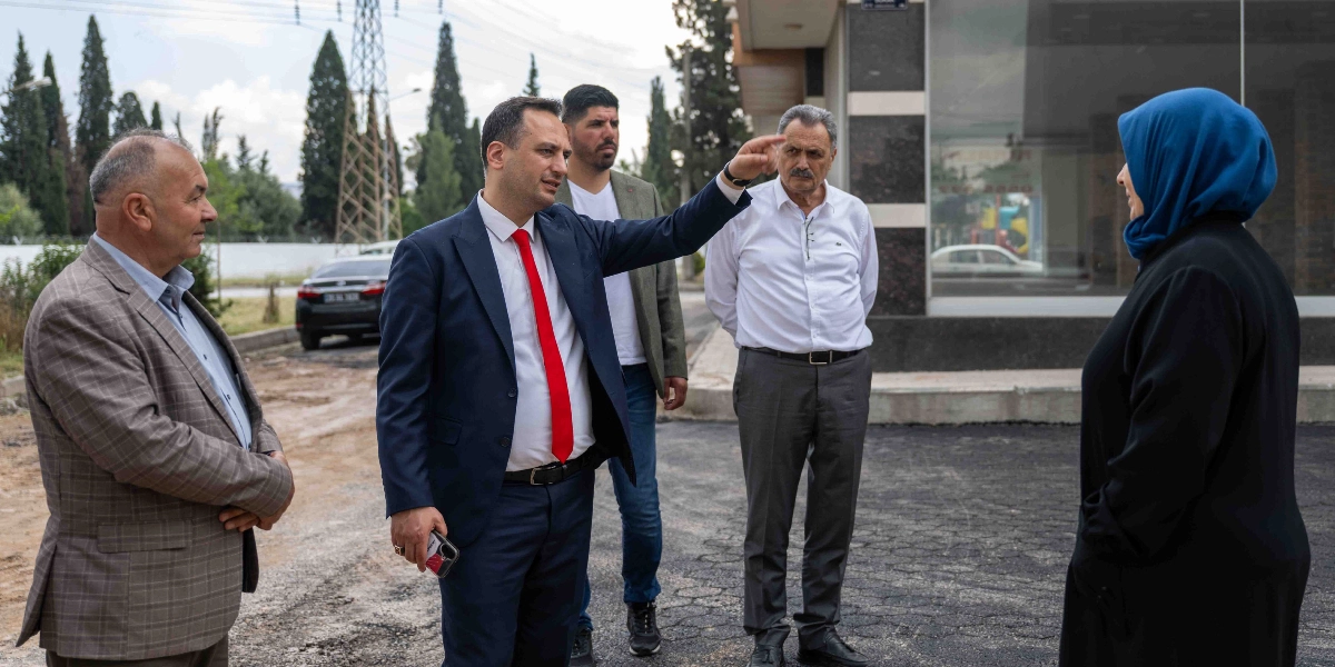 Koyu renk takım elbiseli ve kırmızı kravatlı bir yetkili, yeni asfaltlanmış bir yol üzerinde yanında bulunan üç kişiye eliyle bir yönü işaret ederek bilgi veriyor. Yanındaki grupta ekose ceketli bir beyefendi, beyaz gömlekli bir beyefendi ve mavi başörtülü bir kadın yer alıyor. Arka planda ise ağaçlar, park halindeki bir otomobil ve modern bir bina girişi bulunuyor.