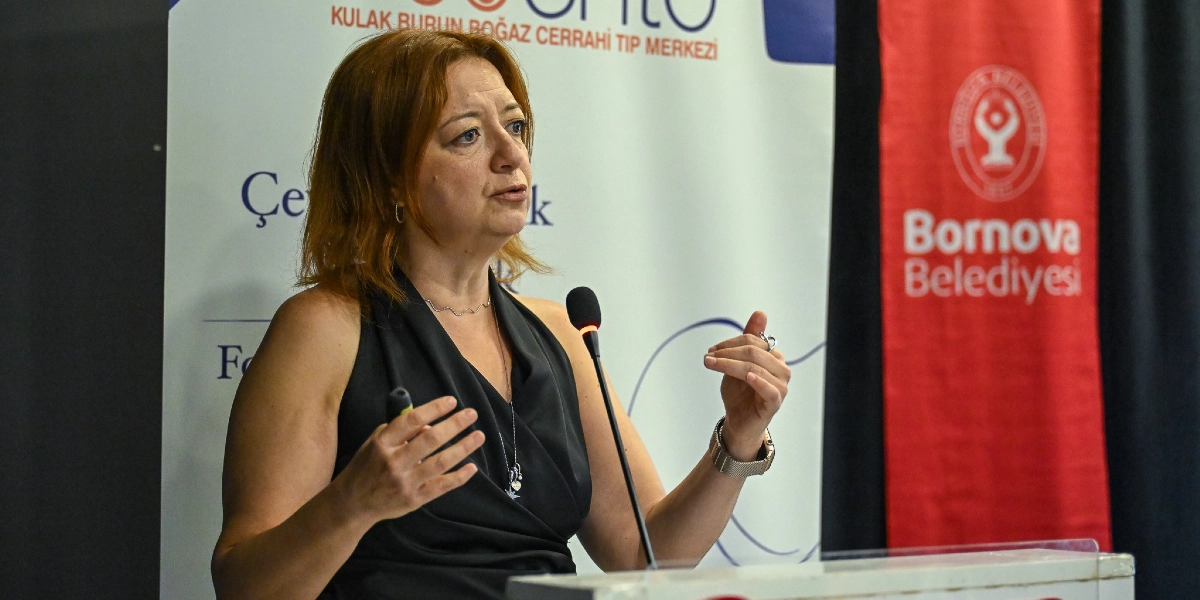 Kızıl saçlı, siyah kolsuz bir bluz giymiş bir kadın, bir kürsüde mikrofon önünde konuşma yapıyor. Arka planda 