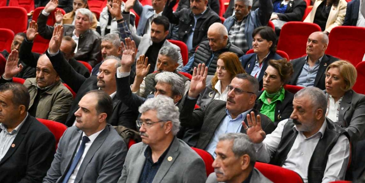 Konferans salonundaki kırmızı koltuklarda oturan bir grup muhtar, bir oylama veya birlik mesajı vermek amacıyla ellerini havaya kaldırmış durumda. Farklı yaş ve giyim tarzlarına sahip kadın ve erkeklerden oluşan kalabalık grup, dikkatle ön tarafa doğru bakıyor.