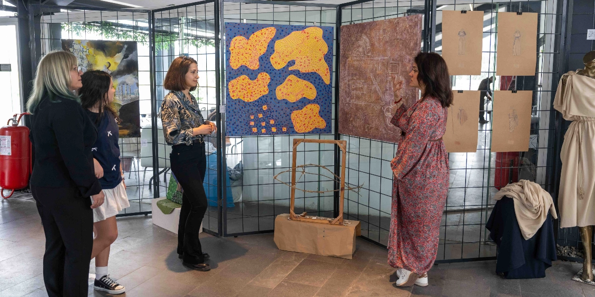 Bornova'daki bir sanat sergisinde, desenli uzun bir elbise giyen kadın, duvardaki eserleri inceleyen üç kişilik bir gruba rehberlik ediyor. Sergide, üzerinde sarı ve turuncu lekeler bulunan mavi bir tablo, antik bir mimari planı andıran dokulu bir eser ve kağıt üzerine çizilmiş figür çalışmaları yer alıyor. Ayrıca, sergi alanının sağ tarafında antik dönem giysilerini anımsatan bir kıyafetin sergilendiği bir cansız manken bulunuyor.