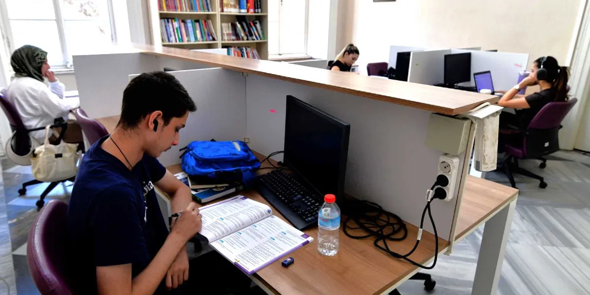 Modern bir kütüphanede, bölmelerle ayrılmış çalışma masalarında ders çalışan öğrenciler. Ön planda, masasında açık bir kitap, bilgisayar ve su şişesi bulunan genç bir erkek öğrenci yer alıyor. Arka planda ise diğer öğrenciler bilgisayarlarında çalışıyor, yan tarafta ise kitaplarla dolu raflar bulunuyor.