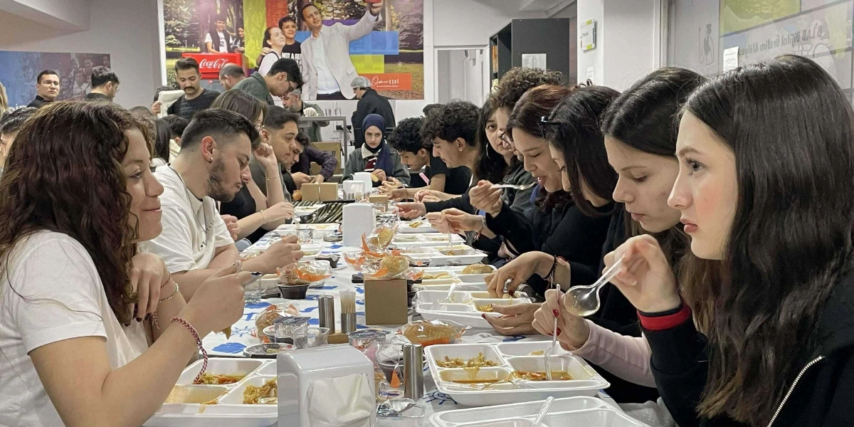 Kapalı bir mekanda, uzun bir masanın etrafında karşılıklı oturmuş gençlerden oluşan bir grup, iftar yemeği yiyor. Beyaz plastik tabldot tabaklarda yemeklerin servis edildiği masada, gençler sohbet ederek yemeklerini yiyor. Arka planda duvarda, üzerinde bir kişinin fotoğrafının ve çeşitli grafiklerin yer aldığı büyük bir afiş bulunuyor.