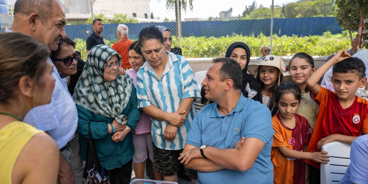 Açık havada, Altındağ Kent Bostanı'nda bir araya gelen farklı yaş gruplarından insanlar, bir masanın etrafında toplanmış sohbet ediyor. Mavi polo yaka tişörtlü bir erkek, kollarını kavuşturmuş şekilde ayakta duran kadınlar ve çocuklarla konuşurken, yanında Galatasaray forması giymiş bir çocuk ve diğer katılımcılar dikkatle onu dinliyor. Arka planda yeşil bitkiler ve tel çitler görülüyor.