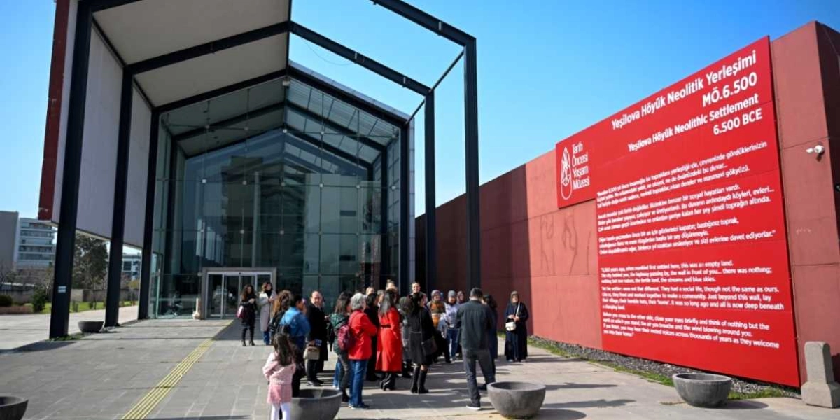 İzmir'deki Yeşilova Höyüğü Neolitik Yerleşimi Müzesi'nin önünde toplanmış bir grup insan görülüyor. Müze binası, modern cam ve çelik mimarisiyle dikkat çekerken, binanın sağ tarafındaki büyük kırmızı duvarda müzenin adı ve tarihsel bilgiler Türkçe ve İngilizce olarak yer alıyor. Müze ziyaretçileri, binanın girişine doğru yönelmiş veya kendi aralarında sohbet ederken görüntüleniyor.