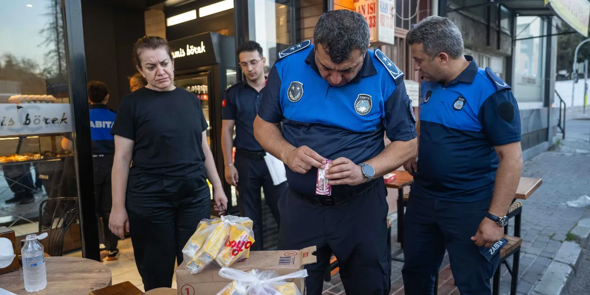 Mavi ve lacivert üniformalı üç zabıta memuru, bir fırın veya börekçinin önünde ürünleri inceliyor. Ortadaki zabıta memuru elindeki paketli bir gıda ürününü dikkatle kontrol ederken, diğer iki memur ona eşlik ediyor. Siyah tişörtlü bir kadın çalışan ise yanında durarak süreci takip ediyor. Önlerinde üzerinde gıda paketleri bulunan bir karton kutu yer alıyor.