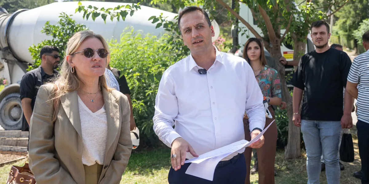 Beyaz gömlekli bir erkek, elindeki belgelerle bir grup insana açıklama yaparken, yanında güneş gözlüklü bir kadın ve arkasında diğer ekip üyeleri yer alıyor. Arka planda ise bir inşaat aracı ve yeşil ağaçlar bulunuyor.