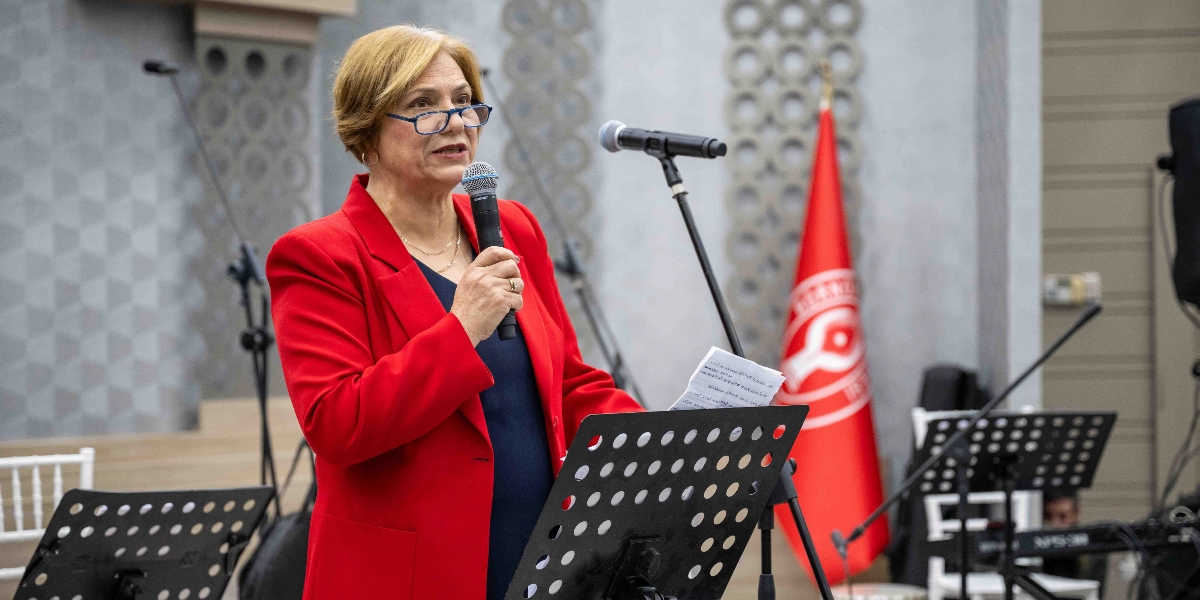 Kısa sarı saçlı, gözlüklü ve kırmızı ceketli bir kadın, elinde mikrofonla bir nota sehpası önünde konuşma yapıyor. Arka planda üzerinde beyaz amblemi bulunan kırmızı bir bayrak ve müzik ekipmanları yer alıyor.