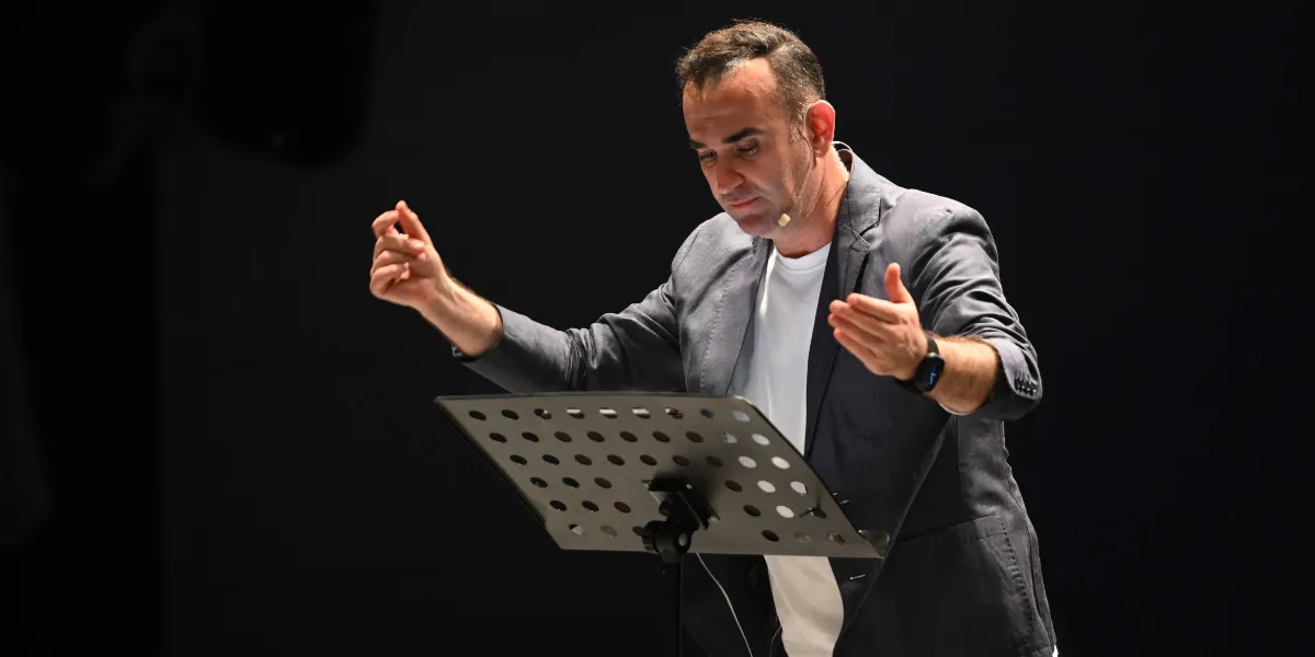 Koyu renkli bir arka plan önünde, beyaz tişört ve gri ceket giymiş bir erkek, önündeki nota sehpasına bakarak elleriyle orkestra şefi gibi hareketler yapıyor.