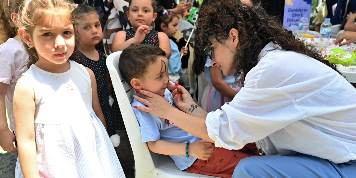 Bornova Büyükpark'ta düzenlenen etkinlikte, kıvırcık saçlı bir kadın, sandalyede oturan küçük bir çocuğun yüzüne boya ile çizim yapıyor. Çocuğun yanında beyaz elbiseli bir kız çocuğu doğrudan kameraya bakıyor, arka planda ise etkinliği izleyen diğer çocuklar yer alıyor. Masanın üzerinde 