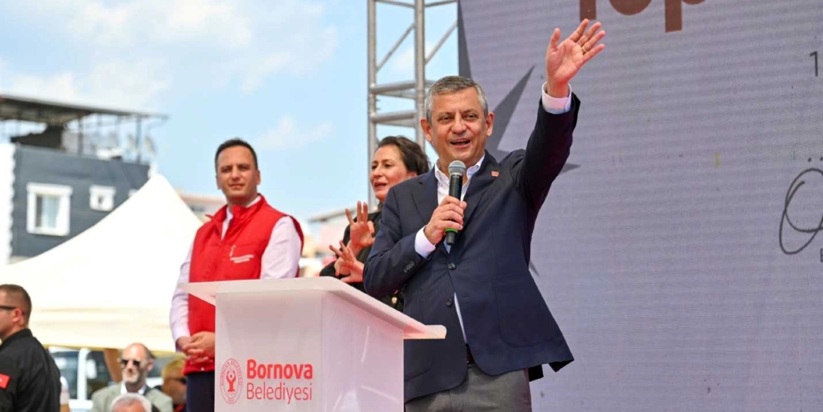 CHP Genel Başkanı Özgür Özel, Bornova Belediyesi logolu bir kürsüde mikrofonla konuşma yaparken sağ elini havaya kaldırarak kalabalığı selamlıyor. Özel'in arkasında kırmızı yelekli bir görevli ve bir kadın yer alıyor. Arka planda ise üzerinde Bornova Belediyesi'ne ait görsellerin bulunduğu büyük bir sahne ekranı bulunuyor.
