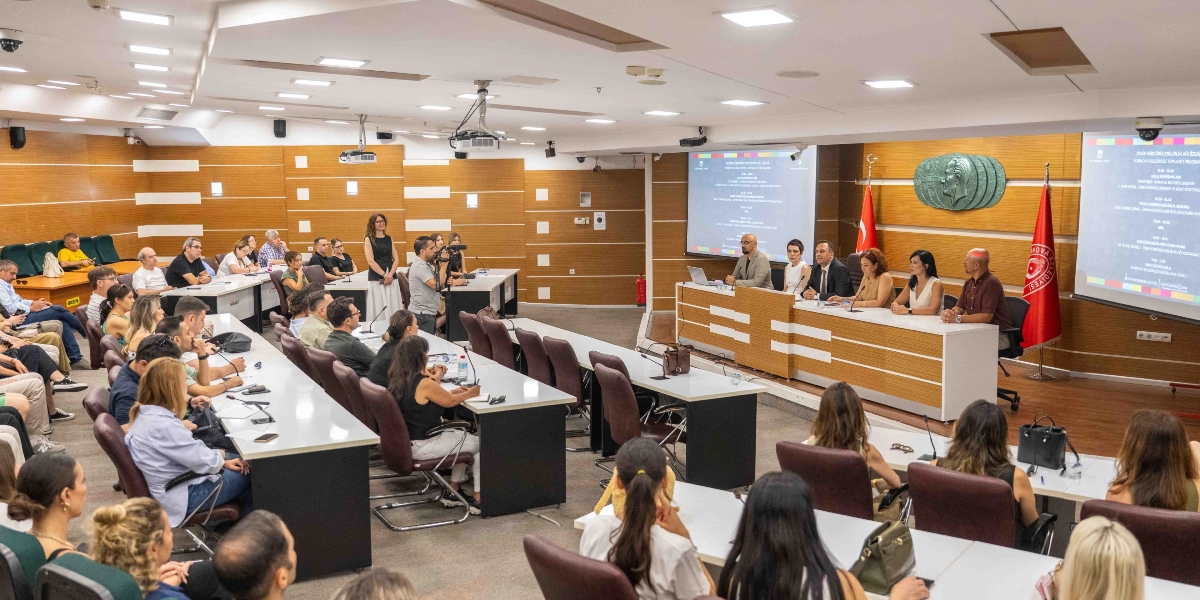 Bornova Belediyesi tarafından düzenlenen, yeşil dönüşüm temalı bir panel veya toplantı salonu. Salonun ön kısmındaki masada beş konuşmacı oturuyor, arkalarındaki duvarda ise Atatürk rölyefi ve iki yanında Türk bayrağı ile Bornova Belediyesi bayrağı bulunuyor. Yan duvarlarda, toplantı programının detaylarının yansıtıldığı iki büyük projeksiyon ekranı yer alıyor. Salonun geri kalanında ise katılımcılar sıralar halinde oturarak konuşmaları dinliyor; bir kişi ise ayakta elindeki kamerayla çekim yapıyor.