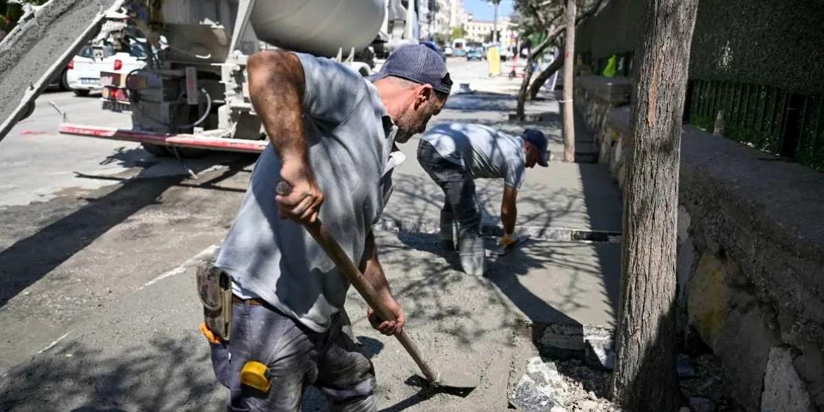 İki işçi, Bornova'da bir kaldırım çalışması sırasında beton mikserinden dökülen taze betonu kürek ve mala yardımıyla düzeltiyor. Ön plandaki işçi kürekle betonu yayarken, arkadaki işçi elindeki malayla yüzeyi pürüzsüzleştiriyor.