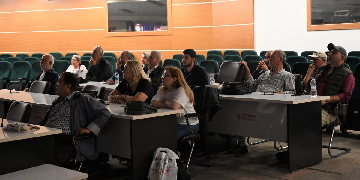 Bornova Belediyesi'ne ait bir konferans salonunda, sıralarda oturmuş bir grup katılımcı dikkatle karşıya bakıyor. Katılımcılar arasında farklı yaş gruplarından kadın ve erkekler bulunuyor; bazıları masalara yerleştirilmiş mikrofonları kullanıyor. Masaların ön yüzünde Bornova Belediyesi logosu yer alıyor. Ön plandaki bir katılımcının sandalyesine asılı beyaz bez çantanın üzerinde 