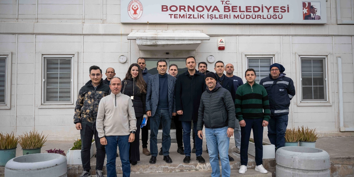 Bornova Belediyesi Temizlik İşleri Müdürlüğü binasının önünde duran, aralarında Bornova Belediye Başkanı Mustafa İduğ'un da bulunduğu bir grup çalışan ve yetkili, objektiflere poz veriyor. Binanın giriş kapısının üzerindeki tabelada T.C. Bornova Belediyesi Temizlik İşleri Müdürlüğü yazısı ve sağ üst köşede Atatürk portresi yer alıyor. Gruptaki kişiler günlük ve iş kıyafetleri içinde, binanın önündeki basamaklarda yan yana dizilmiş şekilde duruyor.