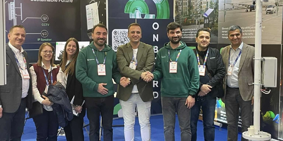 Bir fuar standında yan yana duran yedi kişilik bir grup, yeşil ve dijital dönüşüm odaklı teknolojik çözümlerini sergiliyor. Grubun merkezinde yer alan iki kişi el sıkışırken, diğerleri gülümseyerek kameraya bakıyor. Arka planda, üzerinde 