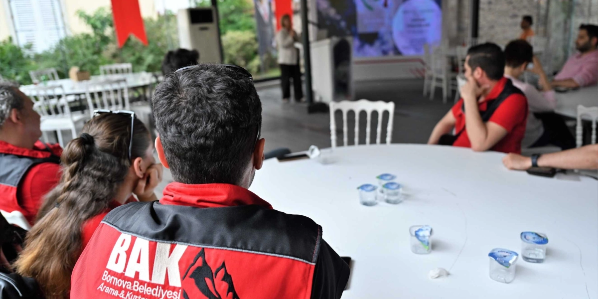 Bornova Belediyesi Arama ve Kurtarma (BAK) ekibi üyeleri, bir eğitim veya bilgilendirme toplantısında masada otururken arkadan görünüyor. Kırmızı siyah yelek giyen ekip üyelerinin önündeki beyaz masada küçük su bardakları bulunuyor. Arka planda ise bir konuşmacı kürsüde sunum yapıyor.