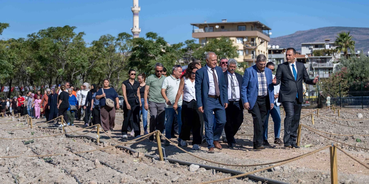 Açık havada, güneşli bir günde, Bornova Belediye Başkanı Mustafa İduğ ve beraberindeki heyet, yeni açılan Kent Bostanı alanında yürüyerek incelemelerde bulunuyor. Toprak zeminli bostan alanında, ahşap kazıklar ve iplerle çevrelenmiş ekim parselleri yer alıyor. Arka planda ağaçlar, bir cami minaresi ve yerleşim yerleri görülürken, heyeti takip eden kalabalık bir grup vatandaş da yürüyüşe eşlik ediyor.