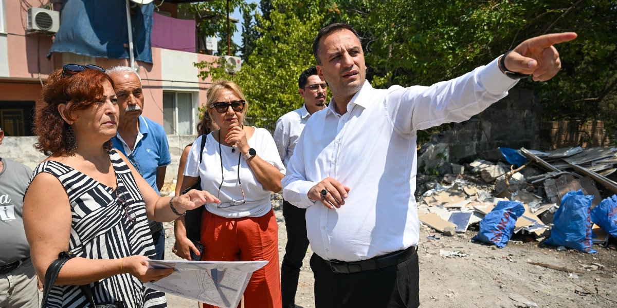Beyaz gömlekli bir erkek, elinde bir proje haritası tutan ve konuşan bir kadın ile yanındaki diğer kişilere, inşaat atıklarının bulunduğu bir alanı eliyle işaret ederek bir şeyler anlatıyor.