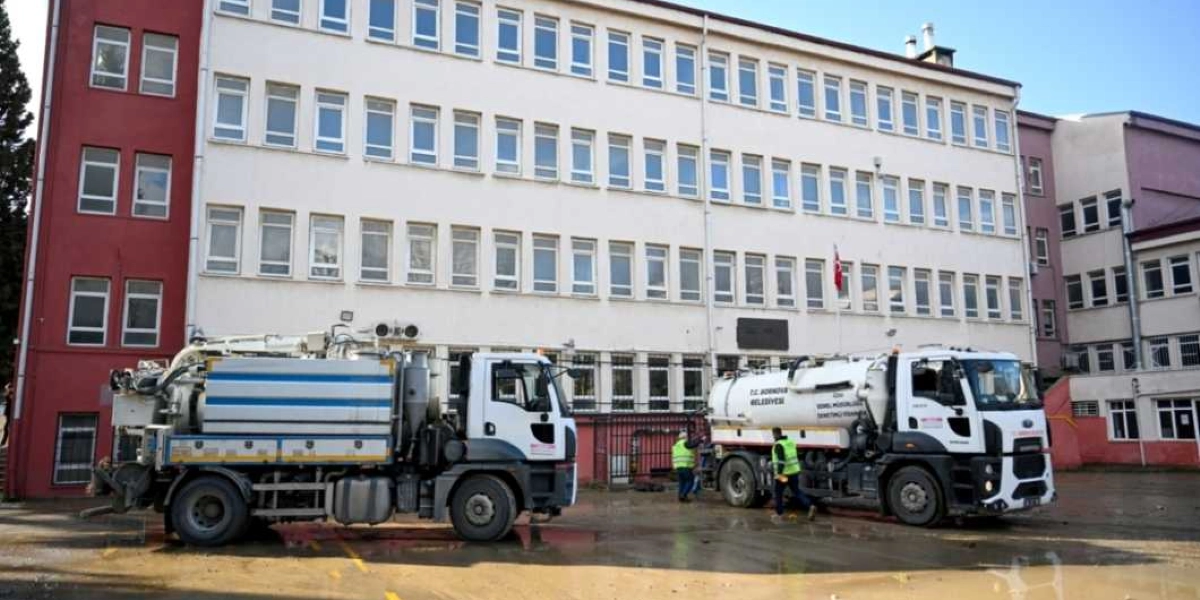 Bornova Belediyesi'ne ait iki adet vidanjör aracı, çok katlı beyaz bir kamu binasının önündeki çamurlu ve su birikintili alanda çalışma yapıyor. Üzerlerinde fosforlu yelek bulunan görevliler araçların yanında duruyor.