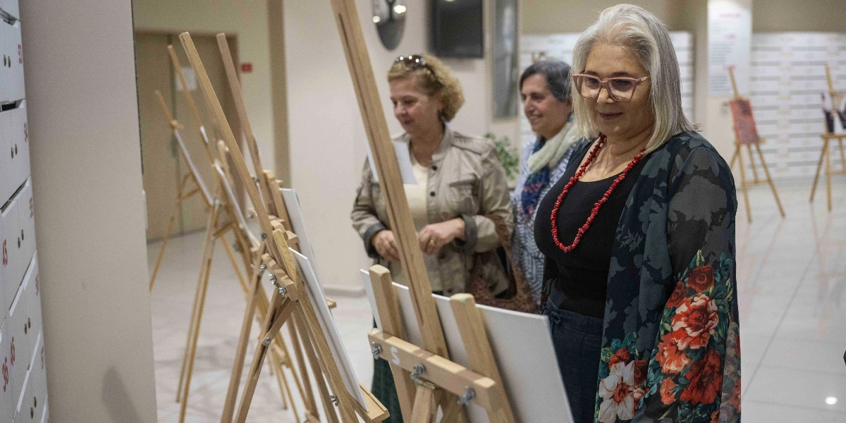 Sanat galerisinde ahşap şövaleler üzerinde sergilenen eserleri inceleyen üç kadın. Ön planda, gri saçlı, gözlüklü, siyah bluz ve çiçek desenli bir hırka giymiş kadın, yanındaki diğer iki kadınla birlikte eserlere bakıyor. Arka planda ise üzerinde numaralar bulunan beyaz dolaplar ve sergi alanının devamında başka şövaleler yer alıyor.
