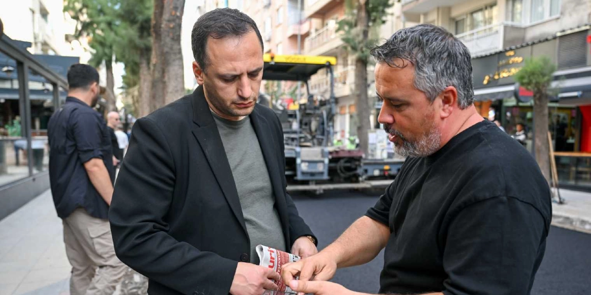 Süvari Caddesi'ndeki yenileme çalışmaları sırasında, siyah ceketli bir yetkili ile siyah tişörtlü bir çalışan, asfalt dökme makinesinin önünde durmuş bir belgeyi inceliyor. Arka planda yol yapım aracı ve caddenin kenarındaki dükkanlar görülüyor.