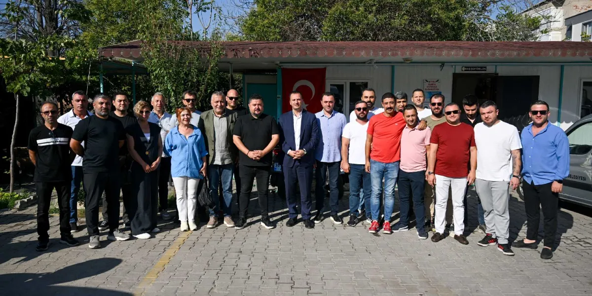 Bornova Belediye Başkanı Ömer Eşki, itfaiye personeli ve belediye çalışanlarıyla birlikte açık havada, bir hizmet binasının önünde toplu halde poz veriyor. Aralarında kadınların da bulunduğu yaklaşık yirmi kişilik grup, gündelik kıyafetlerle yan yana dizilmiş şekilde duruyor. Arka planda Türk bayrağı asılı olan tek katlı prefabrik bir yapı ve ağaçlar yer alıyor.