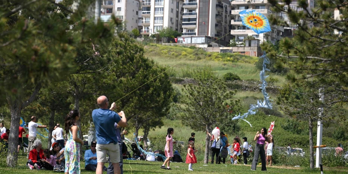 Ağaçlık ve yeşil bir alanda uçurtma uçuran insanlar; ön planda mavi tişörtlü bir adam elindeki ipi tutarak üzerinde güneş figürü olan mavi bir uçurtmayı gökyüzüne doğru yönlendiriyor. Çevrede piknik yapan, yürüyen ve uçurtmalarla oynayan çocuklar ve yetişkinler bulunurken, arka planda ise yamaçta yer alan çok katlı apartmanlar görülüyor.