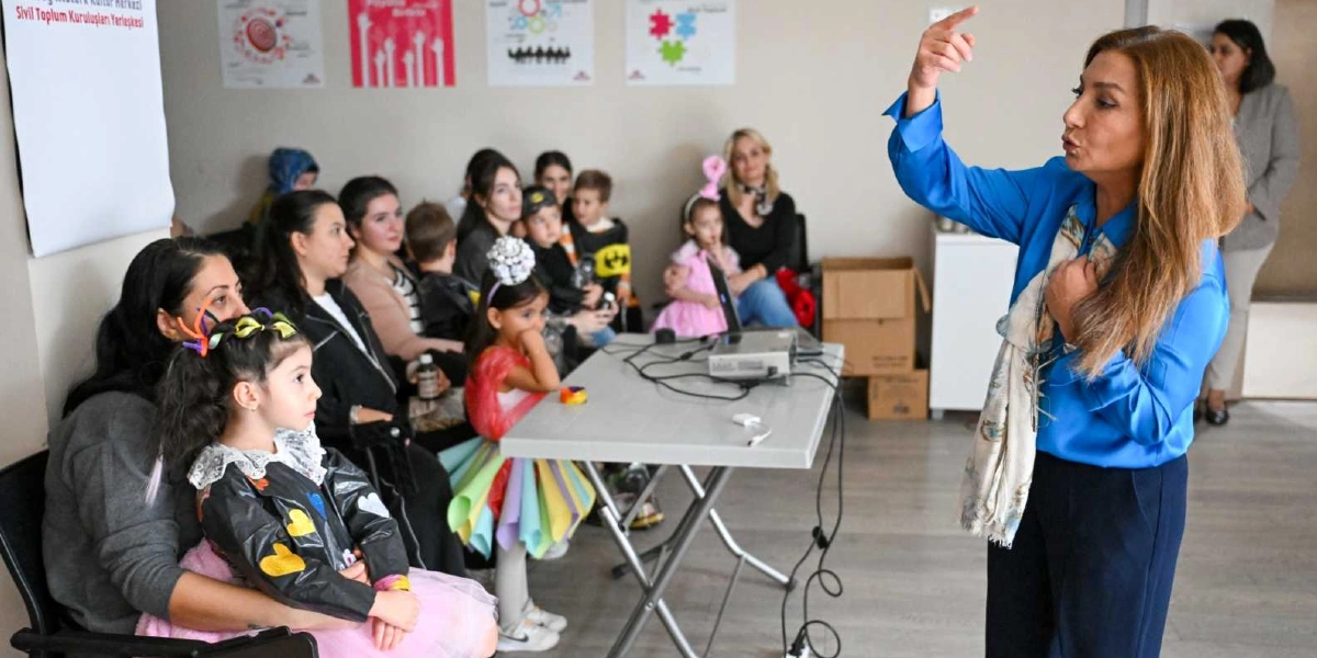 Mavi gömlekli bir kadın, bir sınıfta veya etkinlik alanında çocuklara ve ebeveynlere hitaben konuşma yapıyor. Kadın, el hareketleriyle bir şeyler anlatırken, önünde oturan çocuklar ve yetişkinler dikkatle onu dinliyor. Çocuklardan bazıları renkli ve yaratıcı kostümler giymiş durumda. Arka planda duvarda çeşitli bilgilendirici afişler asılı ve odanın ortasında üzerinde bir dizüstü bilgisayar bulunan bir masa yer alıyor.