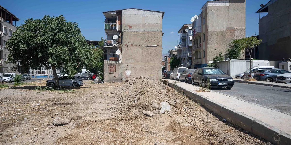 Bornova'da, ön planda toprak ve moloz yığınıyla kaplı boş bir arsa, sol tarafta büyük bir ağaç ve sağ tarafta park halindeki araçların bulunduğu asfalt bir yol yer alıyor. Arsanın arka planında ise dış cephesi sıvasız, çok katlı apartmanlar ve açık mavi bir gökyüzü görülüyor.