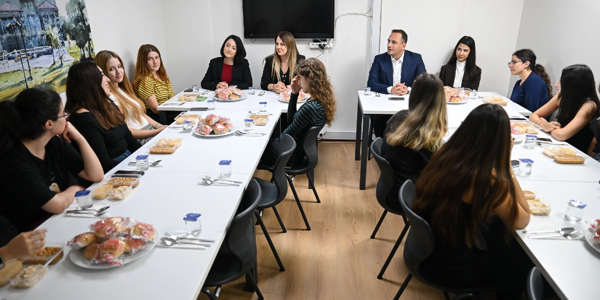 Bornova Belediye Başkanı Ömer Eşki, bir öğrenci yurdunda öğrencilerle bir araya gelerek sohbet ediyor. U-şeklinde birleştirilmiş masaların etrafında oturan öğrenciler ve belediye yetkilileri, önlerinde hazırlanan yemek paketleri ve atıştırmalıklarla birlikte toplantı yapıyor.