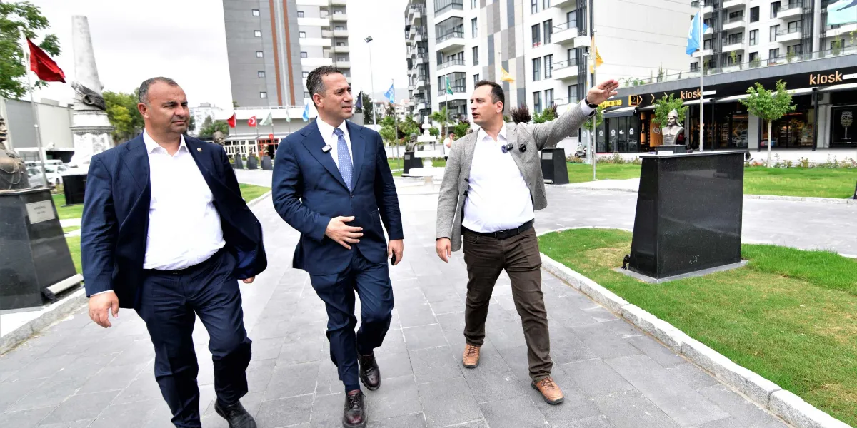 Üç erkek, Bornova Abide-i Hürriyet Meydanı'nda yürürken sohbet ediyor. Soldaki iki kişi lacivert takım elbiseli, sağdaki kişi ise gri ceket ve kahverengi pantolon giyerek eliyle ileriyi işaret ediyor. Arka planda modern binalar, heykeller ve çevre düzenlemesi yapılmış bir park alanı görülüyor.