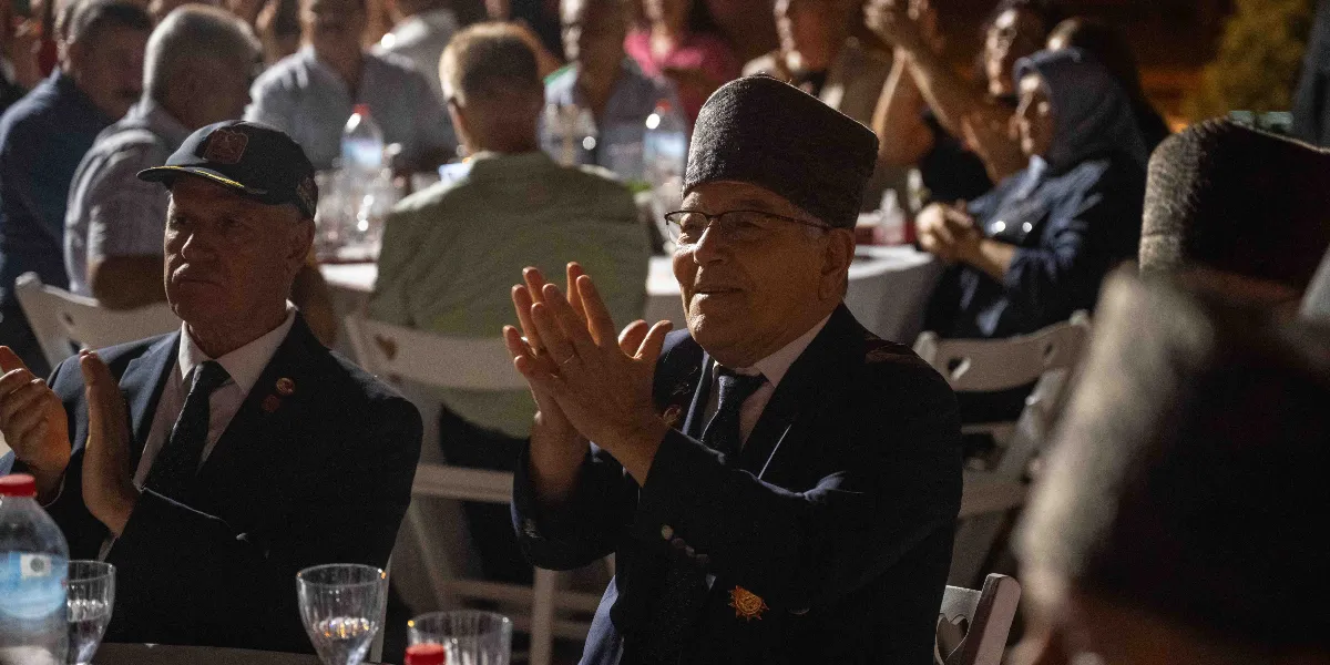 Gece düzenlenen bir etkinlikte, takım elbiseli ve yakalarında madalyalar bulunan yaşlı gaziler masada otururken alkışlıyor. Ön planda, başında kalpak olan ve gözlük takan bir gazi ile yanında lacivert bir şapka takan diğer bir gazi yer alıyor. Arka planda ise diğer katılımcılar masalarda oturuyor.