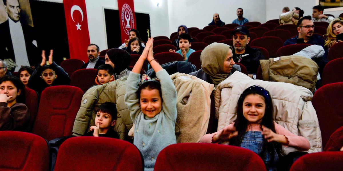 Tiyatro salonunda kırmızı koltuklarda oturan çocuklar ve aileleri, sahnedeki gösteriyi izleyerek alkışlıyor. Ön planda, ellerini havaya kaldırarak neşeyle alkışlayan küçük bir kız çocuğu ve yanında gülümseyen başka bir kız çocuğu yer alıyor. Salonun arka duvarında Atatürk portresi, Türk bayrağı ve Bornova Belediyesi'nin logosu bulunuyor.