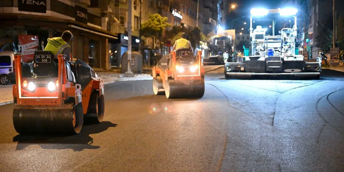 Gece vakti Mustafa Kemal Caddesi'nde devam eden yol yenileme çalışmaları kapsamında, üzerinde sarı yelekli operatörlerin bulunduğu iki adet Hamm marka silindir ve ileride bir asfalt serim makinesi çalışıyor. İş makinelerinin farları yanıyor ve çevredeki dükkanların ışıklarıyla aydınlanan caddede asfalt serme işlemi yapılıyor.