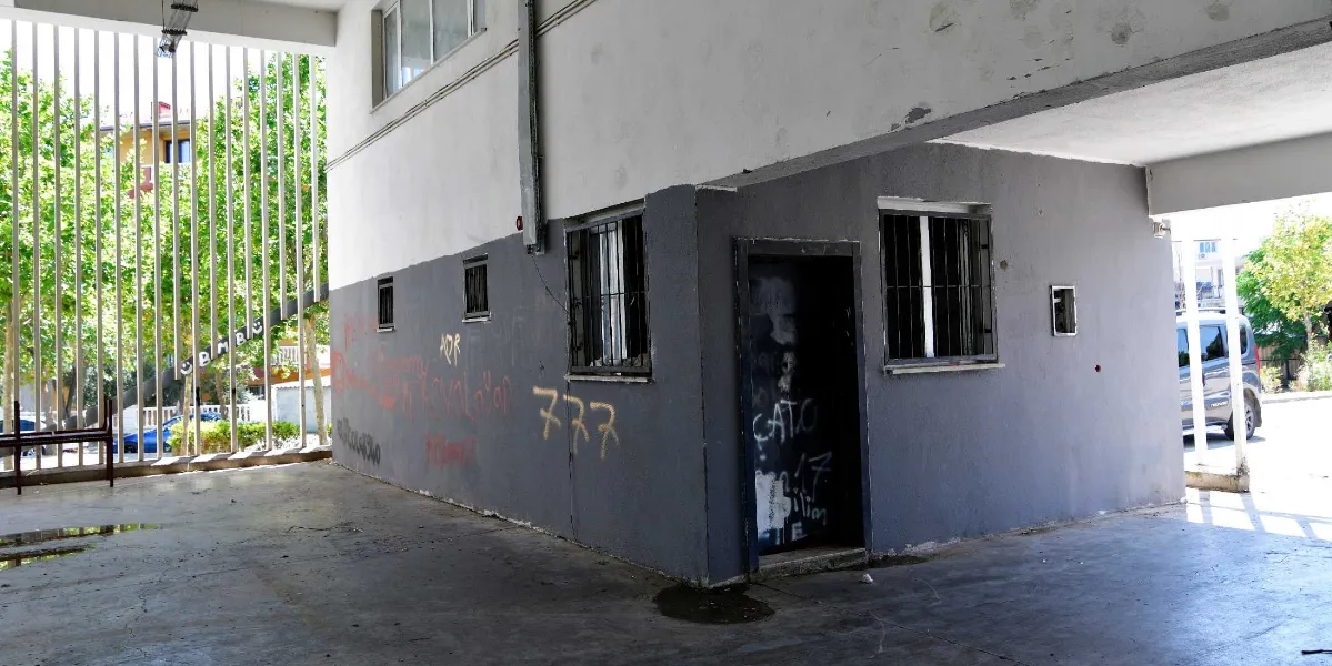 Gri boyalı dış cephesinde çeşitli sprey boya yazıları ve çizimlerin bulunduğu, demir parmaklıklı pencerelere ve açık bir kapı boşluğuna sahip tek katlı bir yapı. Binanın önünde dikey metal çubuklardan oluşan bir çit ve zemin seviyesinde yer yer su birikintileri görülüyor.