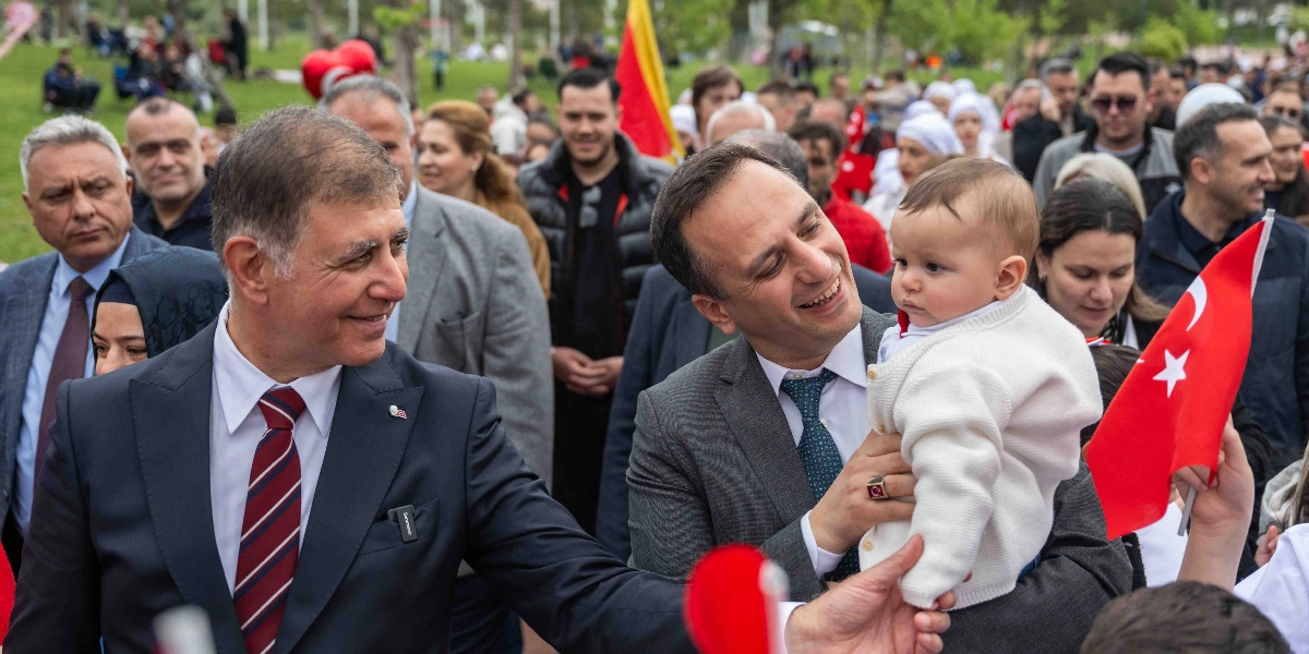 Açık havada, kalabalık bir etkinlik alanında takım elbiseli iki erkek, kucaklarında beyaz hırkalı bir bebekle gülümseyerek yürüyor. Öndeki takım elbiseli ve kırmızı çizgili kravatlı adam gülümseyerek yan tarafa bakarken, yanındaki diğer adam kucağındaki bebeğe sevgiyle bakıyor. Arka planda yeşil bir alan, Türk bayrakları taşıyan insanlar ve etkinlik katılımcıları yer alıyor.