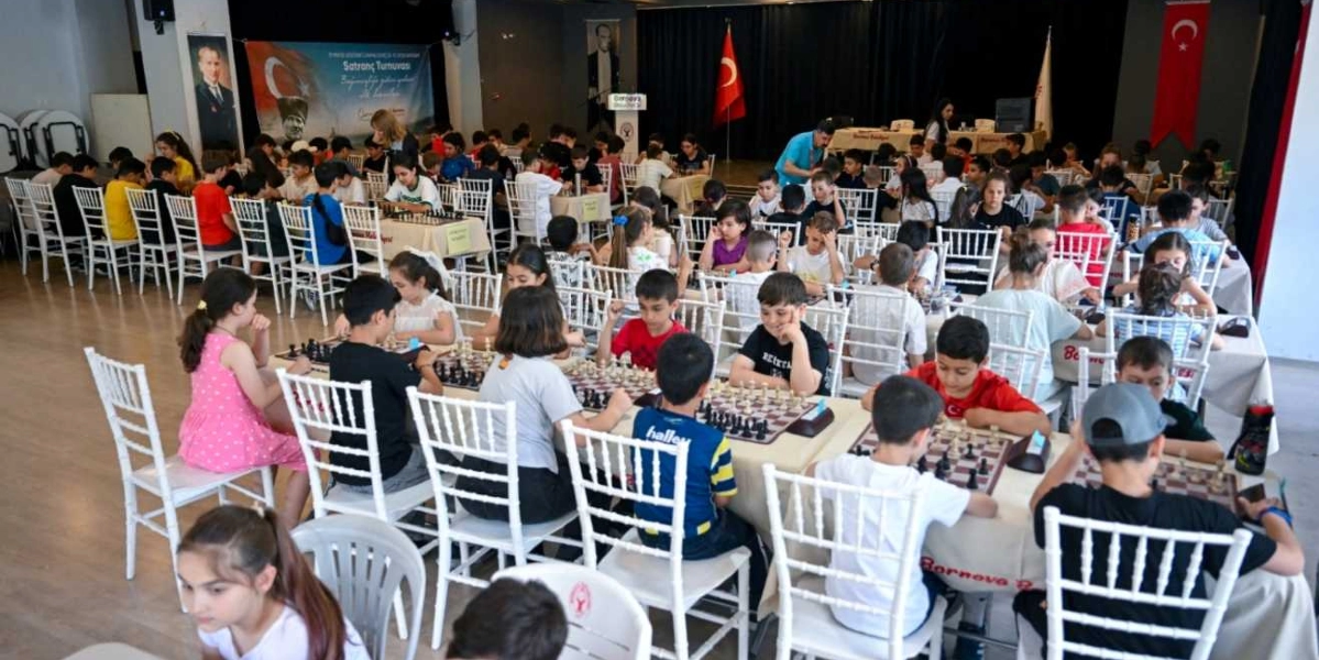 Geniş bir kapalı spor salonunda düzenlenen satranç turnuvasında, beyaz sandalyelerde oturan çok sayıda çocuk ve genç, masalara yerleştirilmiş satranç tahtaları başında oyun oynuyor. Salonun arka planında Türk bayrakları, Atatürk posterleri ve turnuvayı belirten bir afiş yer alıyor. Odaklanmış bir şekilde hamlelerini düşünen katılımcılar, turnuva atmosferinde ciddiyetle satranç mücadelesi veriyor.