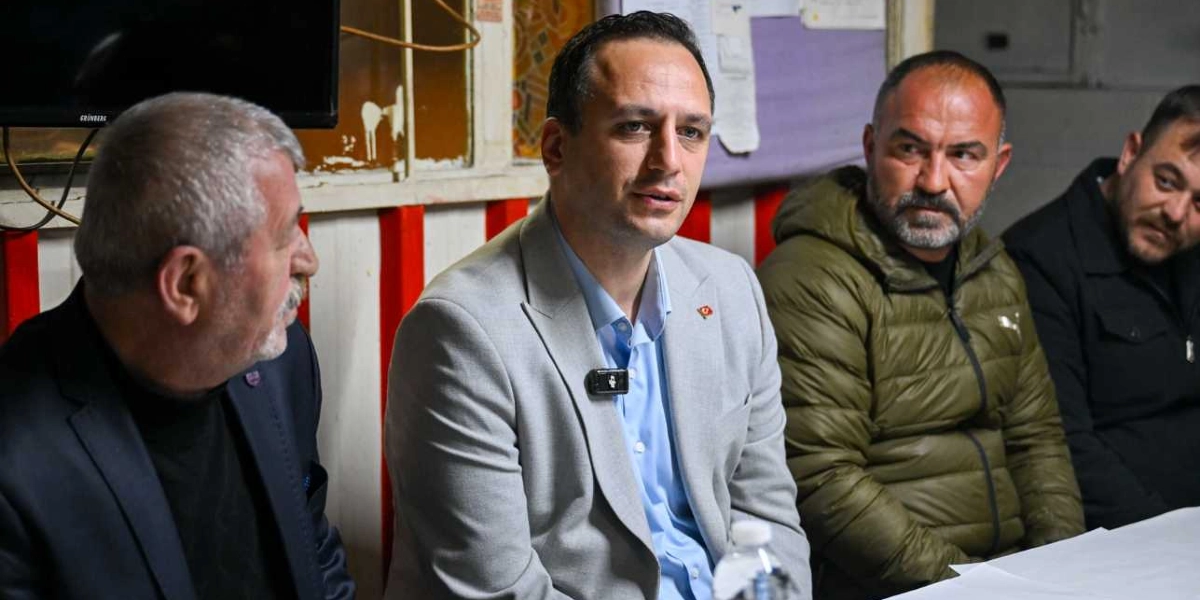 Açık renkli ceket ve mavi gömlek giymiş bir kişi, yanında oturan üç erkekle birlikte bir masada oturarak konuşuyor. Ortadaki kişinin yakasında küçük bir Türk bayrağı rozeti ve yaka mikrofonu bulunuyor. Arka planda kırmızı beyaz dikey çizgili bir duvar ve üzerinde bazı notların asılı olduğu bir pano yer alıyor.
