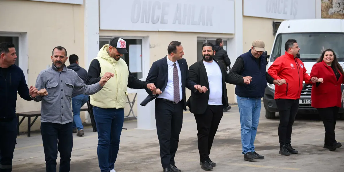Belediye Başkanı Ömer Eşki ve beraberindeki bir grup, açık havada el ele tutuşarak halay çekiyor. Arka planda üzerinde 