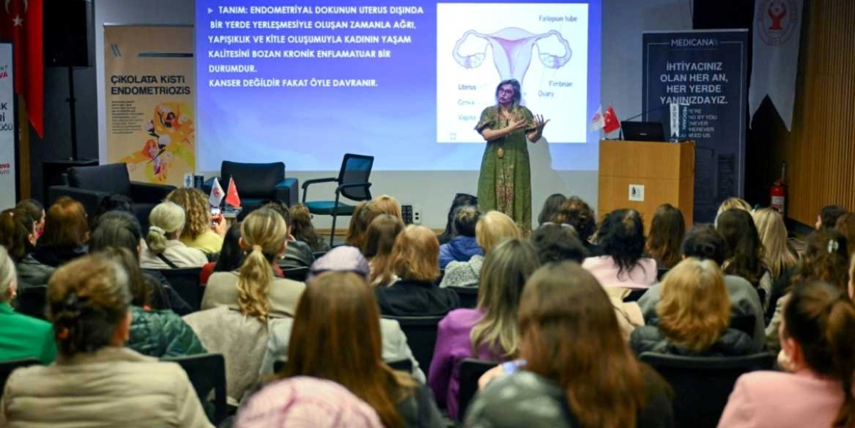 Bir konferans salonunda, kürsüde duran bir konuşmacı, arkasındaki dev ekranda endometriozis hastalığının tanımını ve kadın üreme sistemini gösteren tıbbi bir şemayı yansıtarak katılımcılara sunum yapıyor. Dinleyiciler arasında ağırlıklı olarak kadınların bulunduğu bir grup, konuşmacıyı dikkatle dinliyor. Sahnenin yanlarında 