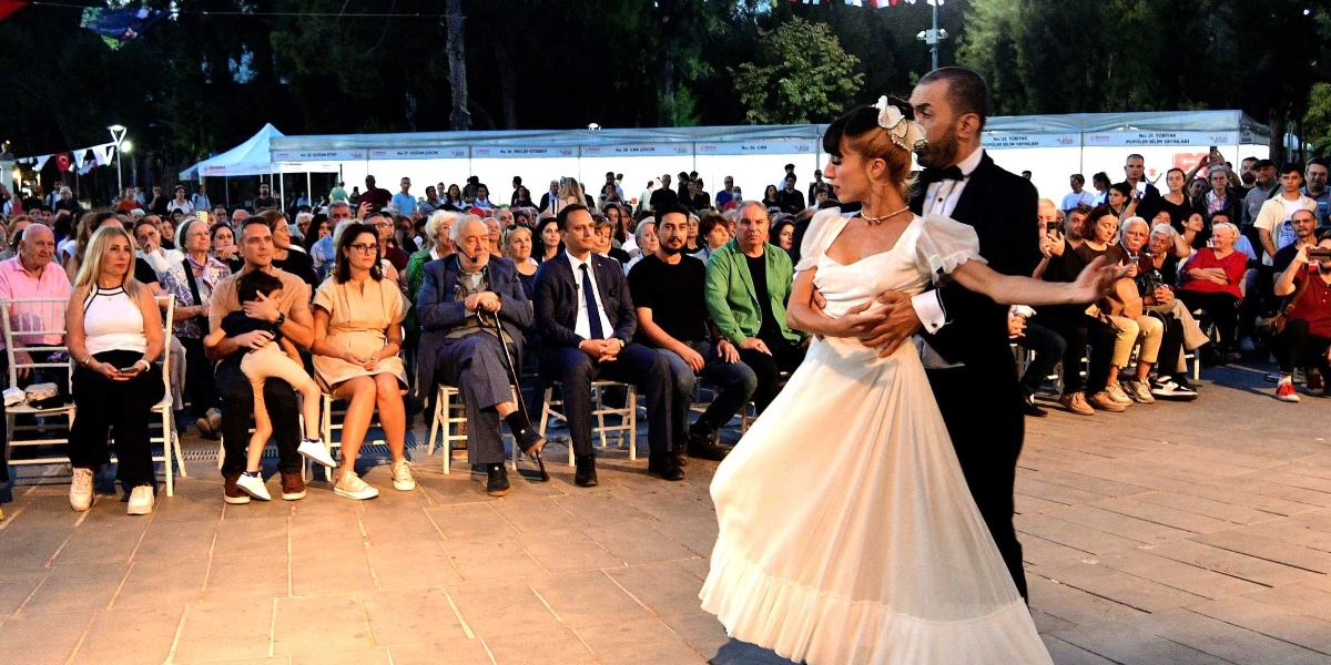 Açık havada düzenlenen bir etkinlikte, beyaz uzun bir elbise giymiş kadın dansçı ile siyah takım elbiseli erkek partneri dans ederken, arkalarında oturan kalabalık izleyici grubu onları izliyor. İzleyicilerin arasında, elinde bastonuyla oturan tarihçi İlber Ortaylı ve yanında takım elbiseli yetkililer dikkat çekiyor. Arka planda ise beyaz çadır stantların bulunduğu bir kitap fuarı alanı görülüyor.