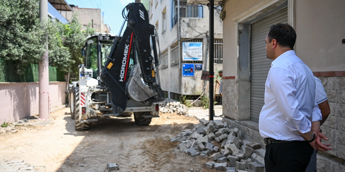 Beyaz gömlekli bir erkek, Çamdibi'nde devam eden yol yenileme çalışmalarını izliyor. Arka planda Hidromek marka bir iş makinesi, sökülmüş parke taşlarının bulunduğu toprak bir zeminde duruyor.