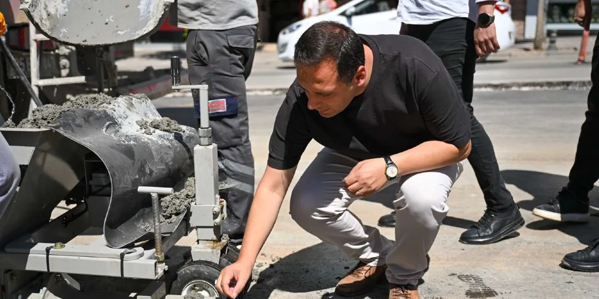 Siyah tişörtlü ve açık renk pantolonlu bir erkek, sokakta beton dökme işlemi yapan küçük, tekerlekli bir makineyi inceliyor. Yanında iş kıyafetli bir çalışan ve diğer kişiler bulunuyor.