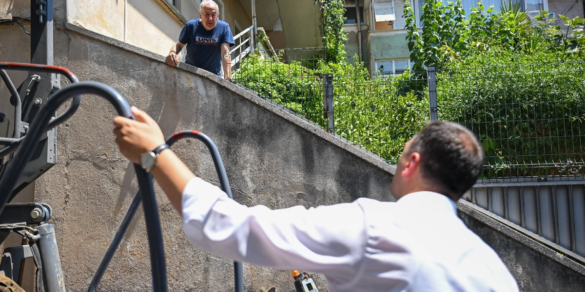 Beyaz gömlekli bir erkek, bir iş makinesinin merdiveninden tutunarak yukarıda, beton bir duvarın arkasında duran yaşlı bir adamla sohbet ediyor.