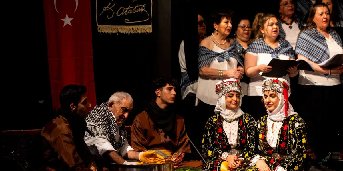 Sahnede geleneksel kıyafetler içinde oturan bir grup insan yer alıyor. Ön planda, yöresel kıyafetli iki genç kadın ve yanlarında oturan, elinde sarı eldivenle bir kapta yemek hazırlayan yaşlı bir adam ile diğer erkekler bulunuyor. Arka planda ise beyaz bluzlarının üzerine mavi ekose şallar takmış, ellerinde notalarla şarkı söyleyen kadınlardan oluşan bir koro yer alıyor. Sahnenin arka duvarında Türk bayrağı ve üzerinde Mustafa Kemal Atatürk'ün imzasının bulunduğu siyah bir pano asılı.