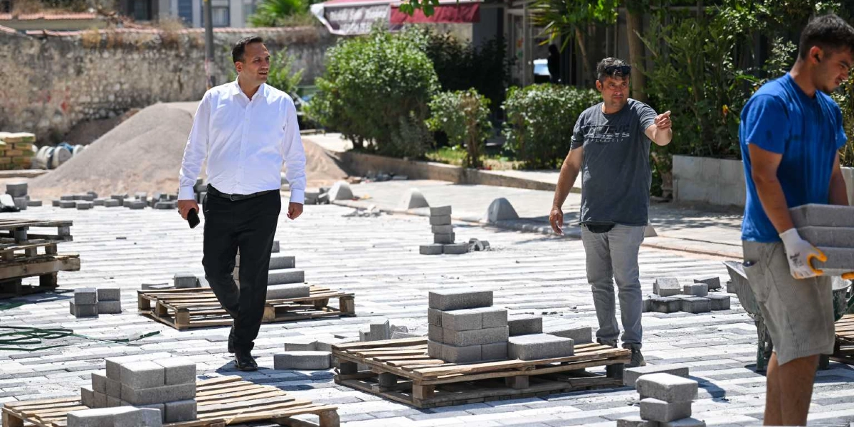 Beyaz gömlekli ve siyah pantolonlu bir yetkili, devam eden bir kaldırım döşeme çalışmasını yerinde inceliyor. Yanında duran gri tişörtlü bir kişi eliyle alanı işaret ederek bilgi verirken, sağ tarafta mavi tişörtlü bir işçi elindeki parke taşlarını taşıyor. Zemin, gri ve beyaz parke taşlarıyla döşenmiş durumda ve çevrede taş yüklü ahşap paletler bulunuyor.